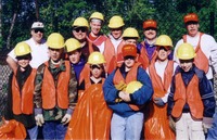 14 scouts and adults smiling at the camera.  Most are wearing orage vests and yellow hard hats.