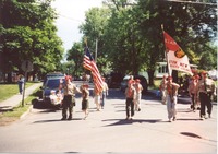 A group of people in BSA class A uniforms marching in the street.  Towards the left, a scout is holding the US flag.  To the right, a scout is holding the Troop 53 flag.