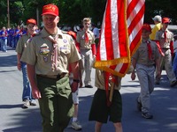 A group of people in BSA class A uniforms marching in the street.  One of whom is holding the US flag, and its blocking their face.