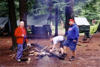 Two adults stand on either side of a scout, who is bent over sawing a log with a bow saw.