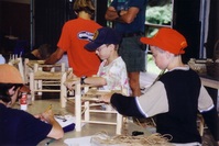 Two scouts weaving baskets.  The one in the middle is wearing a blue hat, the one on the right is wearing the red troop hat.  There's a scout on the left writing something on a clipboard.