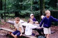 5 scouts sit around a picnic table.  Four are facing to the right, one is facing to the left.