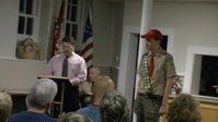 Two people standing in front of the room.  The person on the left if at a podium, reading from something on it; they wearing a pink shirt and tie.  The person on the right is wearing a Class A.