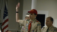 Scout in a class A uniform has his right hand up in the scout sign.  He is speaking.