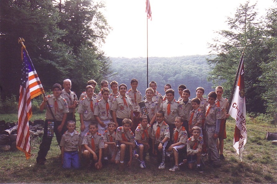 27 people in class A uniforms smiling for the camera in front of an American flag pole.  To the left is a scout holding the US flag, to the right is another scout holding the Troop flag.