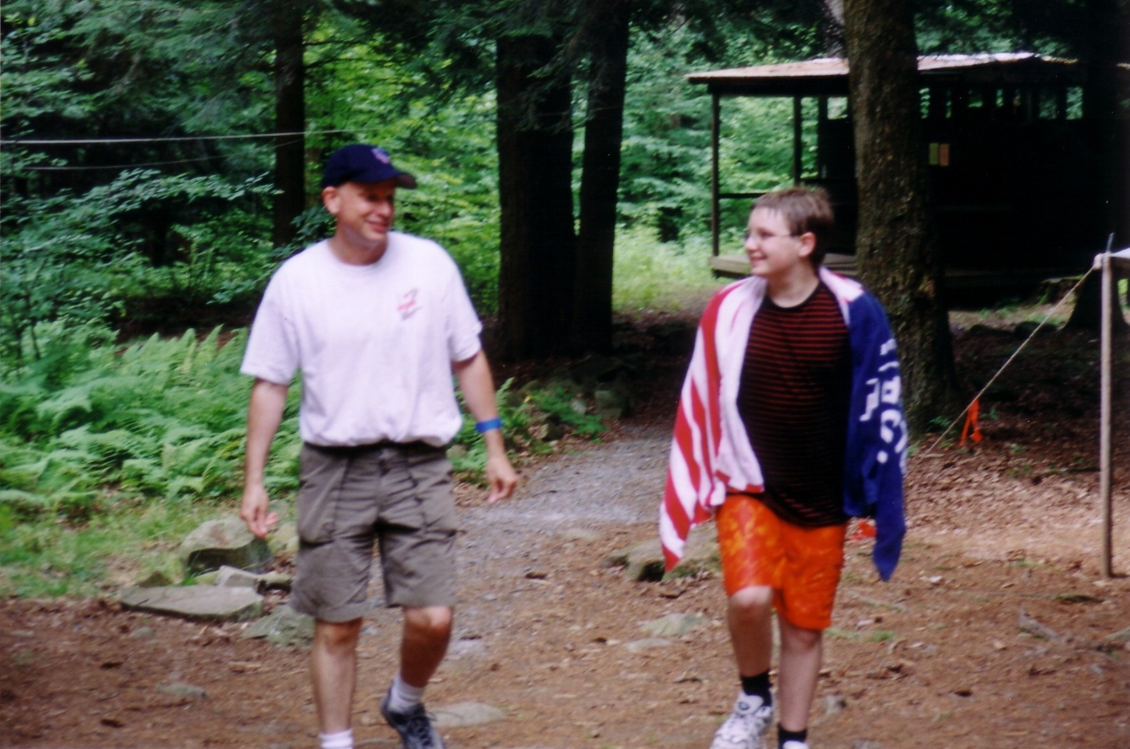 Two people walking towards the camera talking to each other. The person on the right is wet, and wrapped in a towel.
