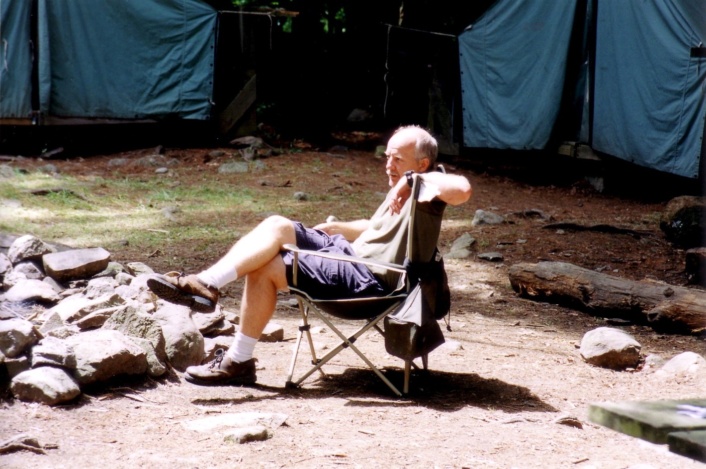A man sitting in a camping chair in the sunlight.  Behind are two cabin tents.
