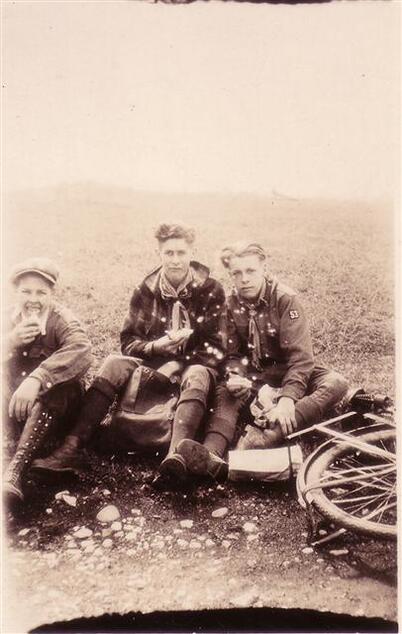 Three scouts in scout uniforms sitting on the ground eating lunch.  There is a bike on the ground to he right of them.