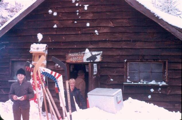 Two scouts standing outside a wooden lodge.  There are several hockey sticks standing up, being stuck in the snow.  A sign says 