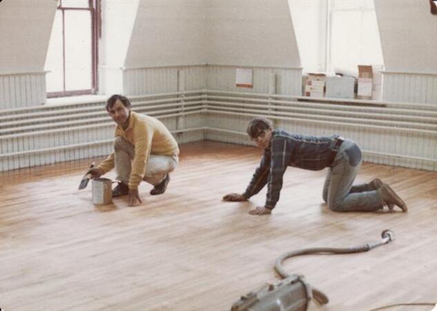 Two people on the floor.  The person on the left is staining the floor, the person on the right appears to be sanding.
