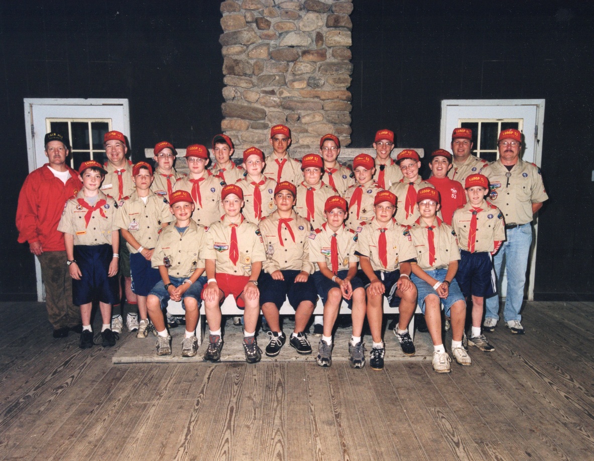 24 scouts and adults posing for a photo in front of a big outdoor fireplace.