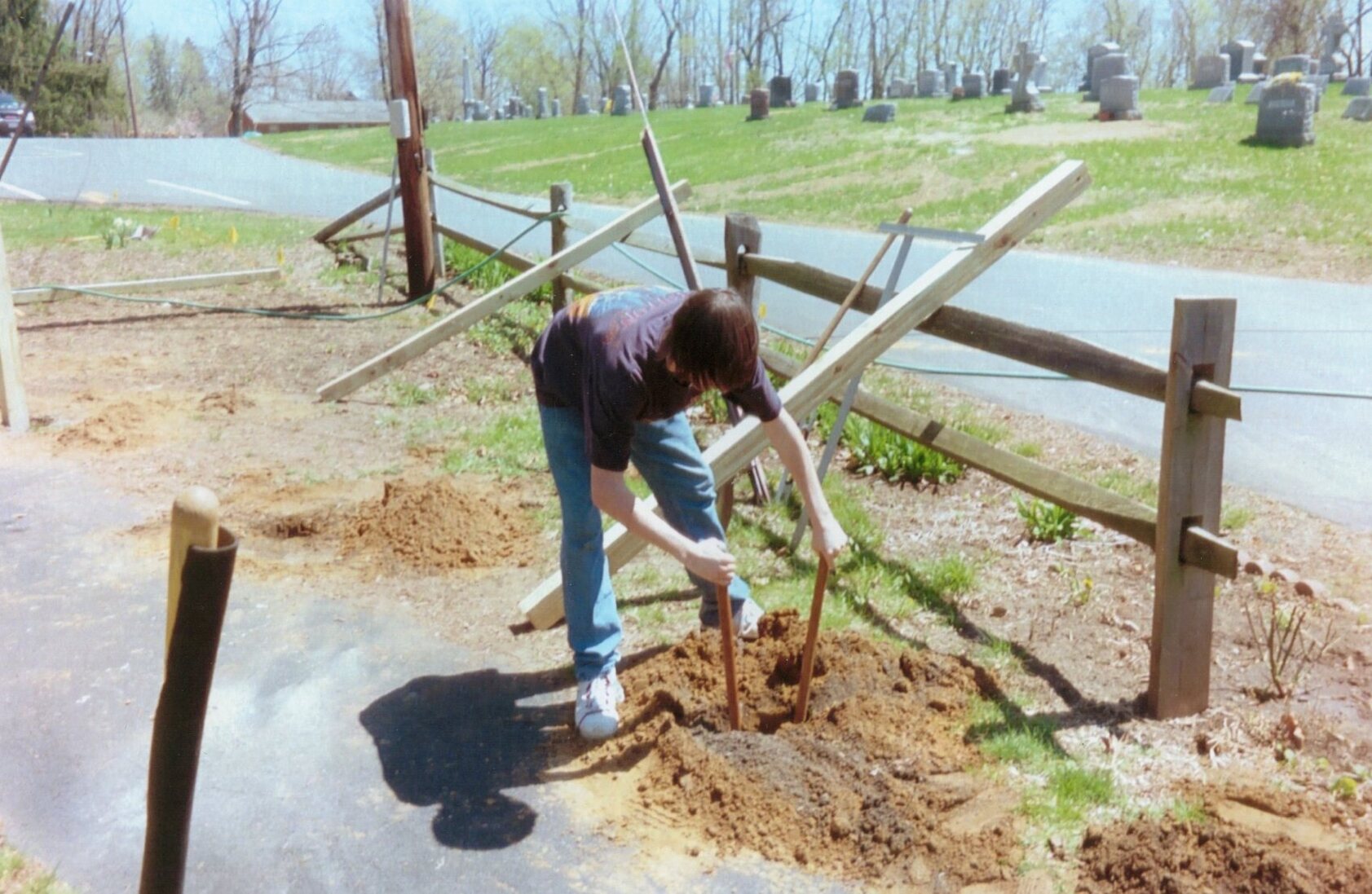 A scout bending down in front of a hole holding onto a post-hole digger inside of the hole.