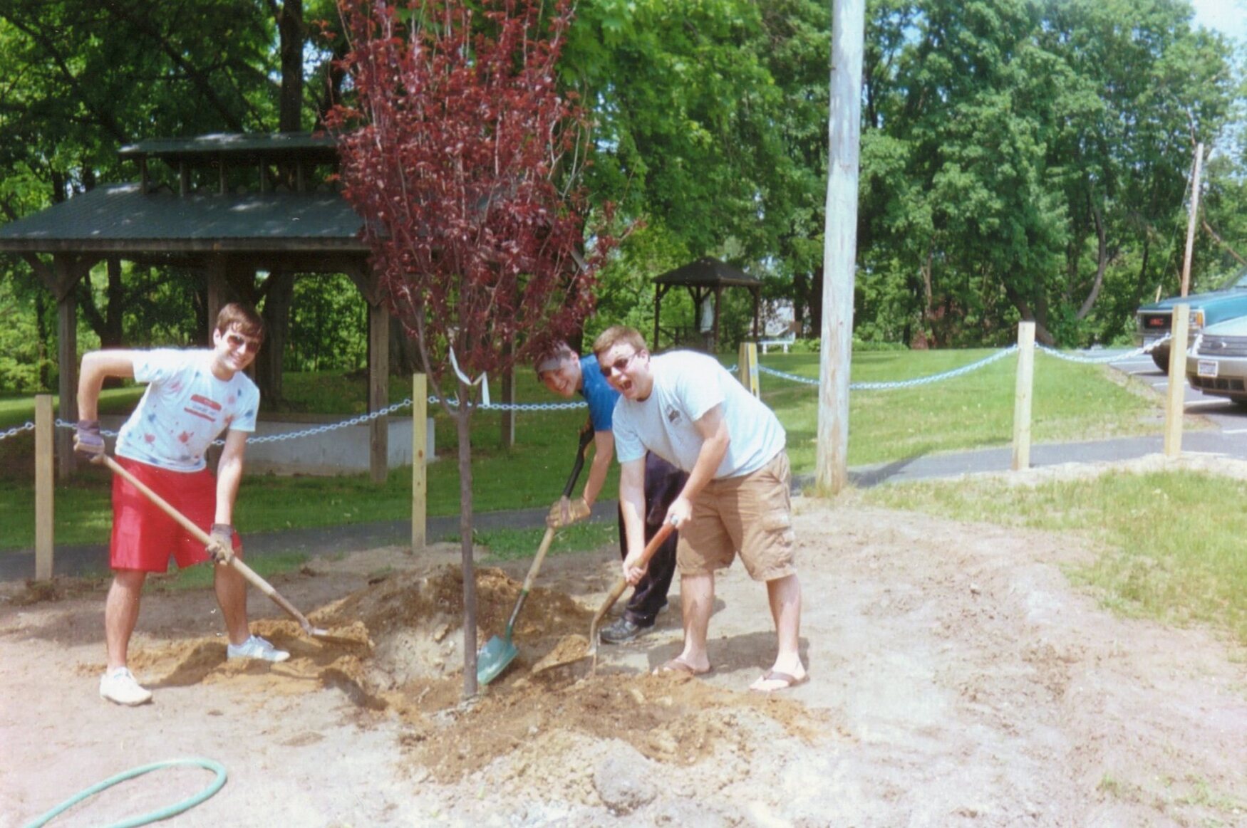 Seth, Brett, and Rob holding shovels filling in a whole that contains a red tree.  They are standing on dirt, and smiling.