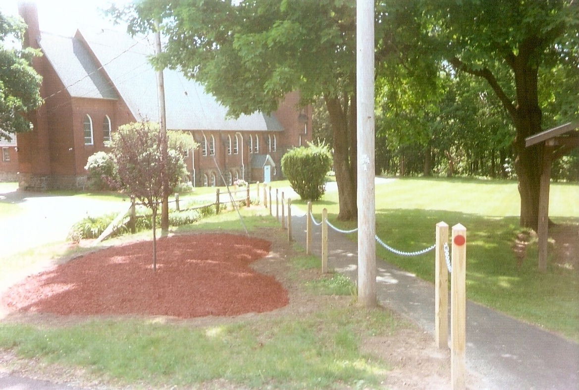 A chain railing to the left of a path.  To the left is a garden with a red tree in it.