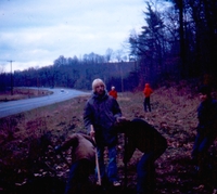 Six people standing in the grass on the side of a two-lane road.  One of them is holding a shovel.