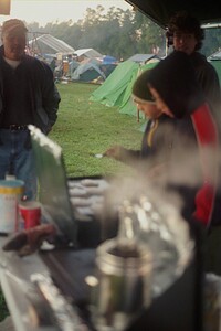 Two scouts are cooking on a grill, while an adult and an older scout observe.
