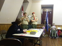2 scouts in class A uniforms standing behind a table.  The scout on the left is holding on to something, while the scout on the right is looking at it.