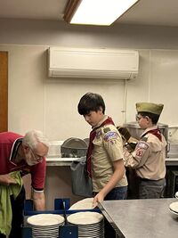 Two scouts washing dishes in Class A uniforms in front on an industrial dishwasher.  There is a man in glasses to their left helping.