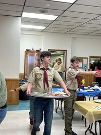 One scout carries a platter of food, while another scout is pointing towards a table.