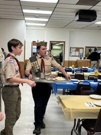 One scout is holding a platter of food, while another one seems to guiding him where to go.