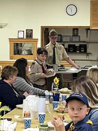 Two scouts serving a table food.  One is holding a tray of food, another is putting a plate on the table.