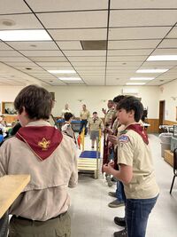 A group of scouts and adult leaders in Class A uniforms are standing around a wooden bridge indoors.  The left side of the bridge is blue, the right side is gold.  There is a single cub scout standing on the blue side, ready to cross over.