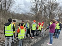 Fourteen people standing along the side of the road.  All but one are wearing yellow reflective vests.