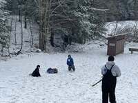 Two scouts are at the bottom of the hill; one is holding a tube, one is sitting on the ground.  Another scout stands looking at them at the top of the hill.