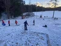 Six scouts sledding down a snowy hill.  There is a frozen lake in the background.
