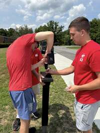 Three people in red shirts drilling a screw into the top of a post.
