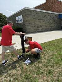 Two people in red shirts screwing a bolt into the bottom of a post.  One person is drilling, the other is holding the post steady.