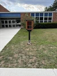 The little library to the right of a sidewalk outside of a brick building.