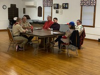 Six people sitting around a table, talking.  Four are wearing Class A uniforms.