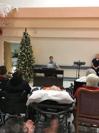 Seth sits behind a piano wearing a bow tie.  There are older folks sitting in the audience.