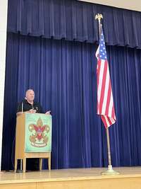 A man wearing a black polo and an eagle metal speaking into a microphone at a podium.