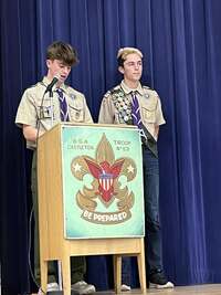 A two scoutsn in tan class A uniforms speaking into a microphone at a podium.