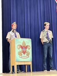 A two scouts in tan class A uniforms speaking into a microphone at a podium.