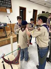 Two scouts are reaching over to help remove the Webelos neckerchief off of a Cub Scout.