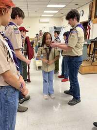 A girl Cub Scout has her Webelos neckerchief removed with the help of an older scout.