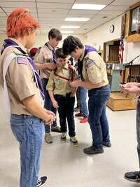 Two older scouts help a younger cub scout put on the Scouts BSA neckerchief.  A scout with red-dyed hair looks towards them.