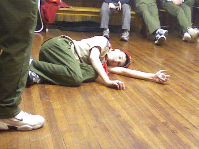 A scout in a class A uniform is lying on the floor on his side, pretending to be hurt.