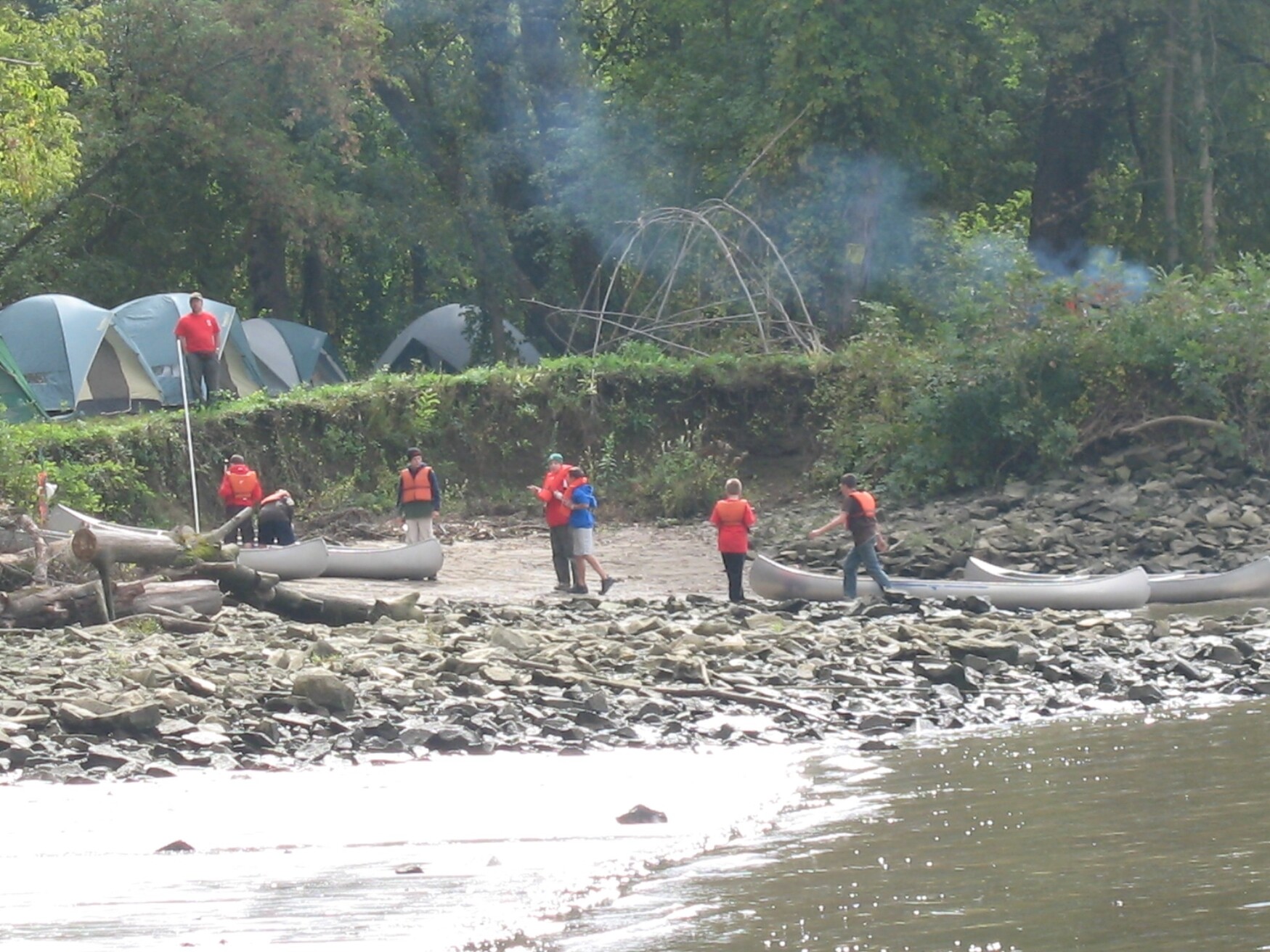 A picture of a rocky beach.  There is a small cliff above the beach, on which there are several tents and the smoke of a fire.
