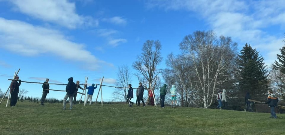 A group of scouts setting up Christmas Tree racks.  The racks are lashed together, and have two A-frame supports on each end, with a thick tree branch going between the two.