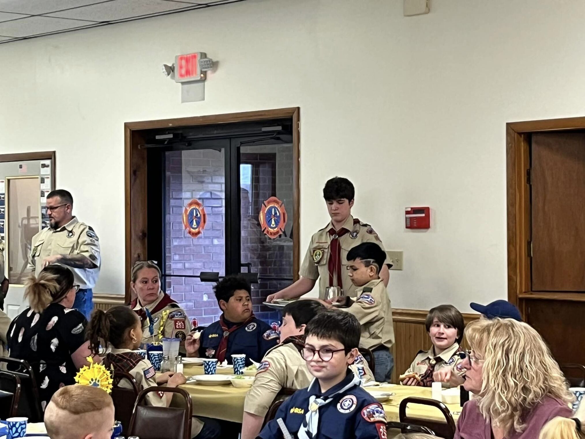 Several people sitting at a table, while two scouts serve them dinner.