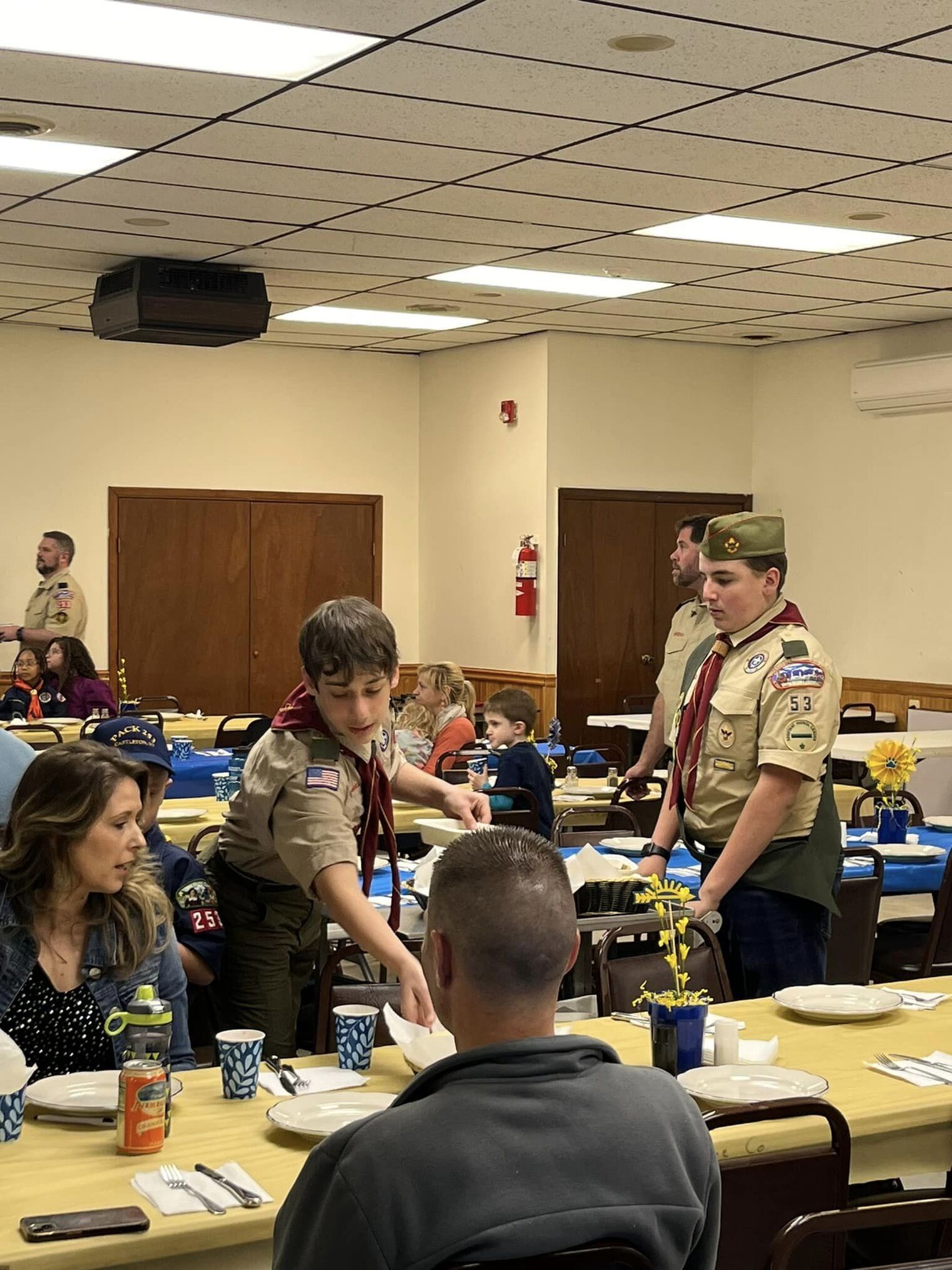 Two scouts serving a table food.  One is holding a tray of food, another is putting a plate on the table.