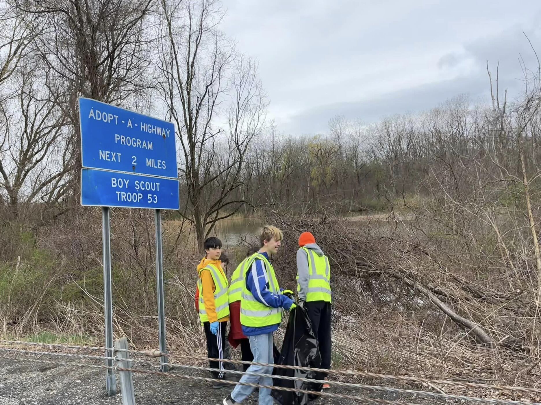 Five scouts cleaning up trash.  Two are hidden being the other three.  All are wearing reflective yellow vests.  There is a sign that says 