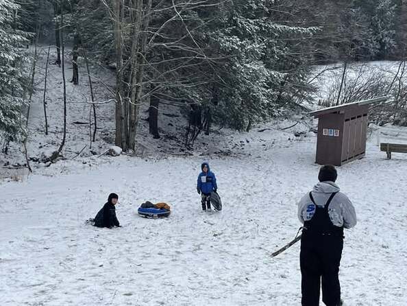 Two scouts are at the bottom of the hill; one is holding a tube, one is sitting on the ground.  Another scout stands looking at them at the top of the hill.