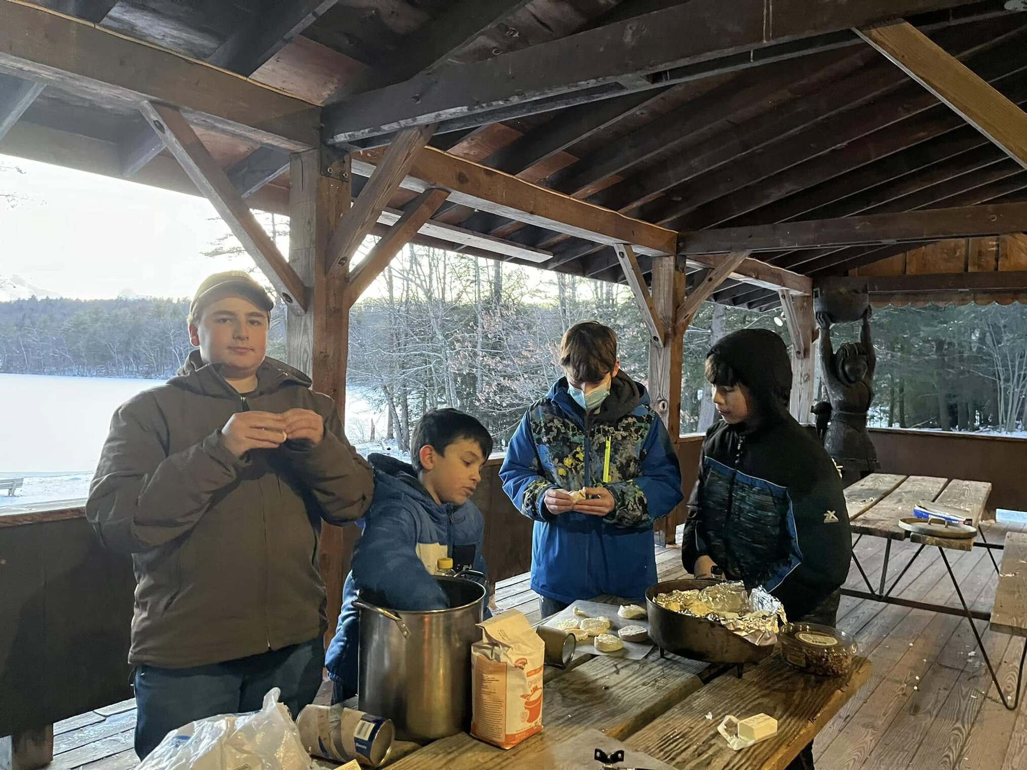 Four scouts prepare a meal.  These are standing around a picnic table with ingredients on the table.