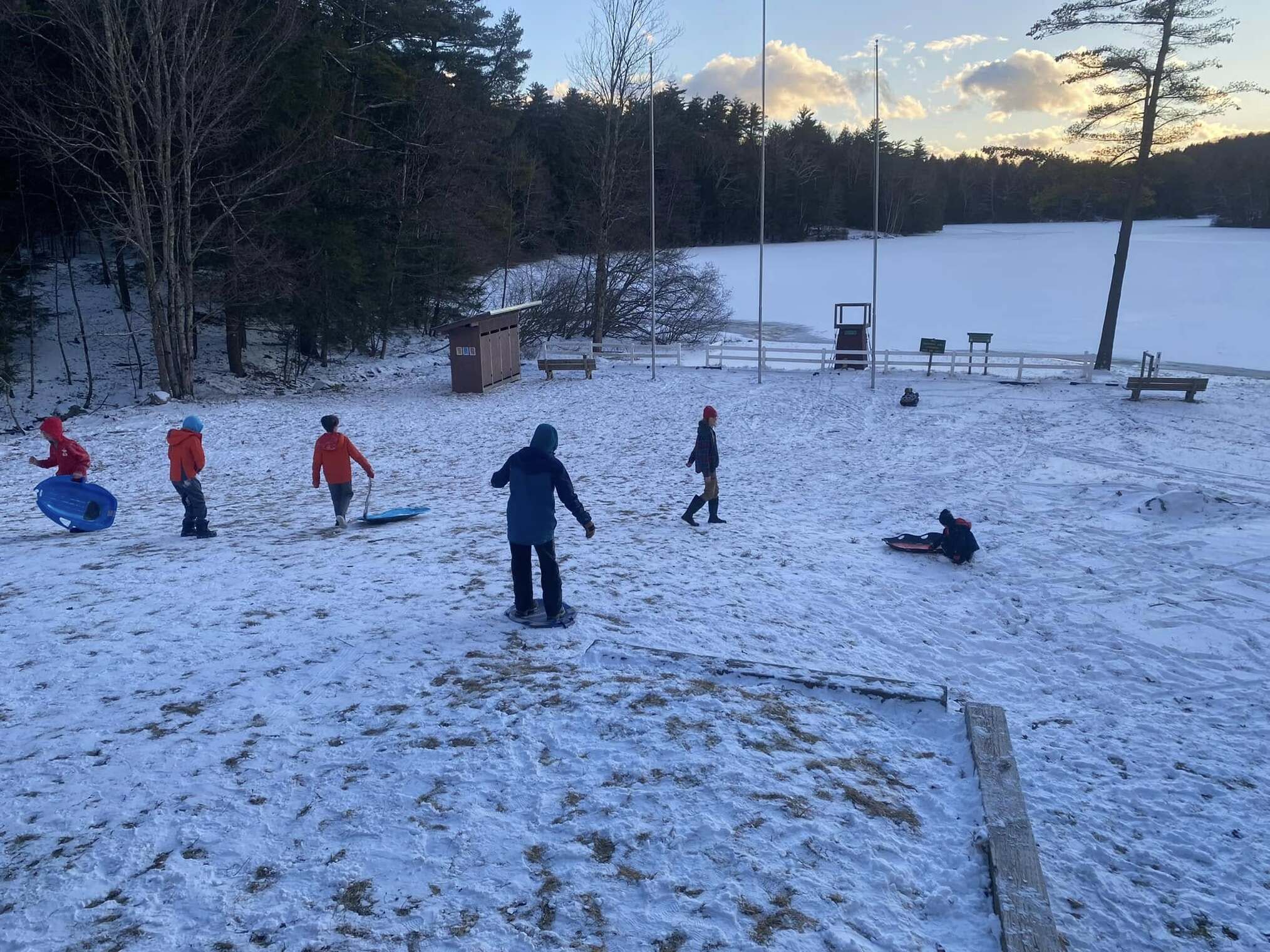 Six scouts sledding down a snowy hill.  There is a frozen lake in the background.