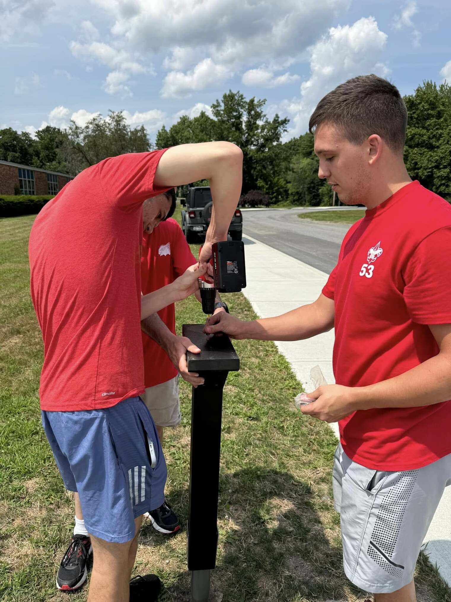 Three people in red shirts drilling a screw into the top of a post.
